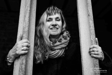 Frau mit langen, blonden Haaren lächelt durch zwei vertikale Stangen hindurch.
