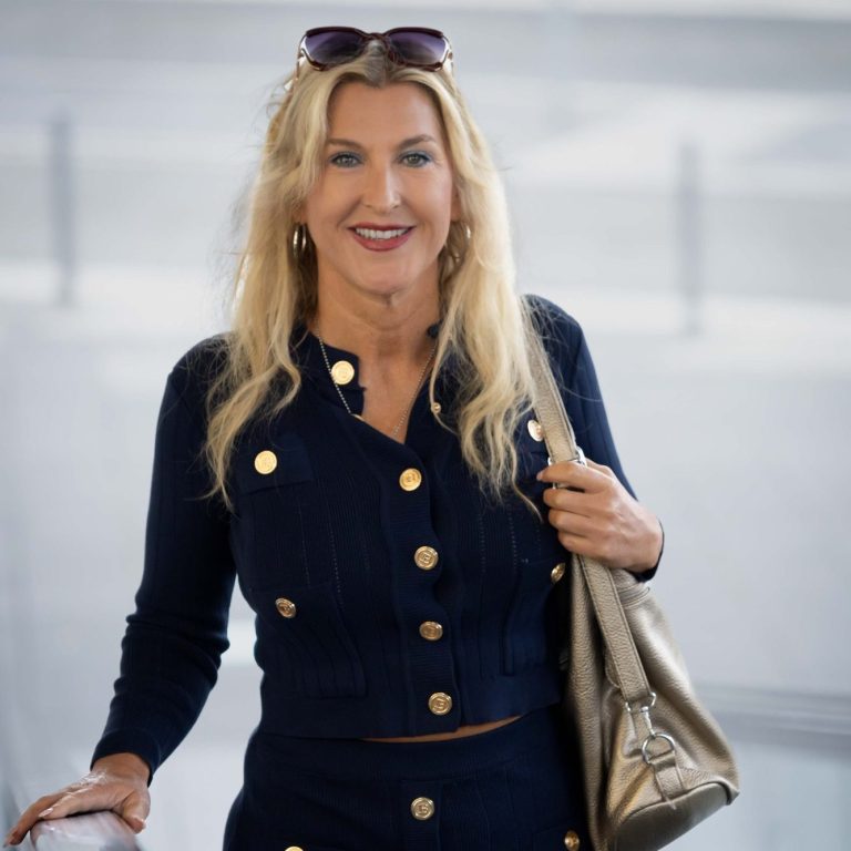 Eine lächelnde Frau mit blonden Haaren trägt eine blaue Jacke und eine silberne Tasche.