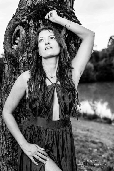 Frau in einem tief ausgeschnittenen Kleid steht neben einem Baum am Ufer eines Sees.