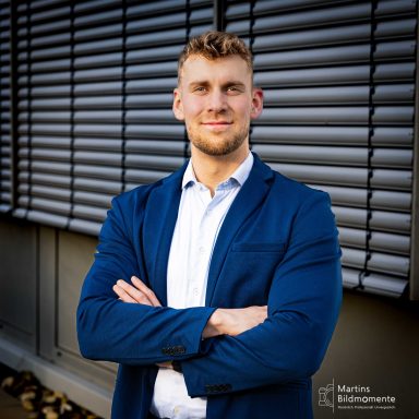 Junger Mann in Blazer, mit verschränkten Armen, vor einer Wand aus Jalousien.