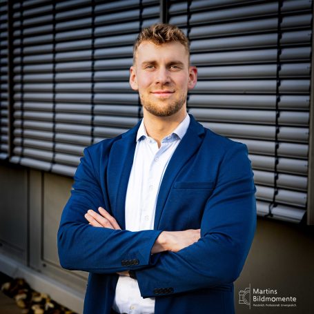 Mann mit grimmigem Ausdruck, Armen verschränkt, trägt einen blauen Anzug vor einer Wand.