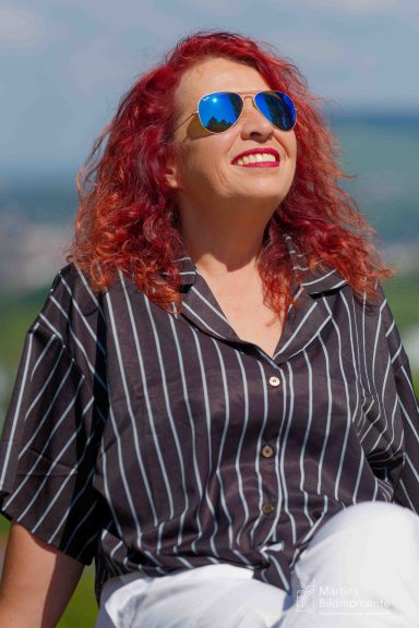Frau mit rot gefärbtem Haar und Sonnenbrille, lächelt unter blauem Himmel.
