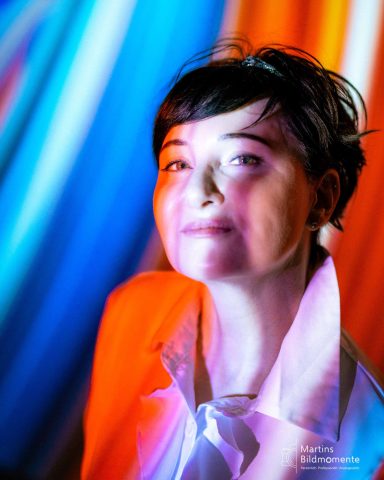 Porträt einer Frau mit kurzem, dunklem Haar vor bunten Lichtstreifen.