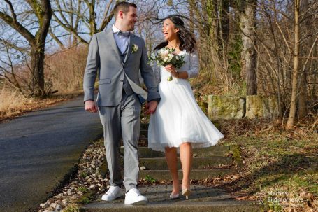 Brautpaar in Hochzeitskleidung spaziert lächelnd auf einem Waldweg.