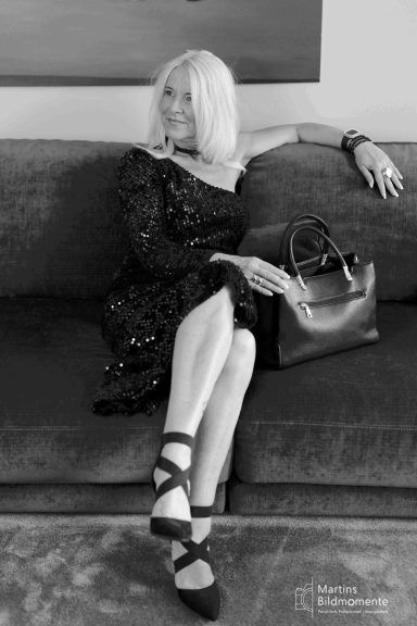 Frau in schwarzem Kleid mit Tasche sitzt entspannt auf einem Sofa.