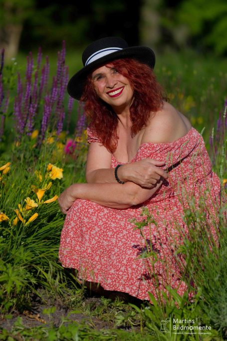 Lächelnde Frau mit rotem Haar in einem Blumenfeld, trägt einen schwarzen Hut und ein rosafarbenes Kleid.