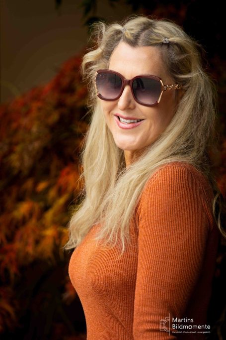 Frau mit langen blonden Haaren und Sonnenbrille, lächelt vor herbstlicher Kulisse.