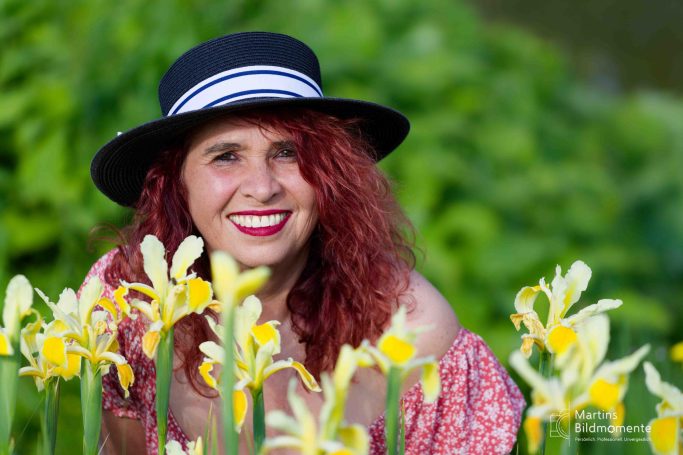 Frau mit roten Locken und Hut, umgeben von gelben Blumen.