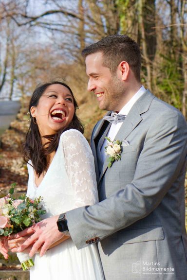 Lächelndes Paar in Hochzeitskleidung, umarmt im Freien, mit Blumenstrauß.