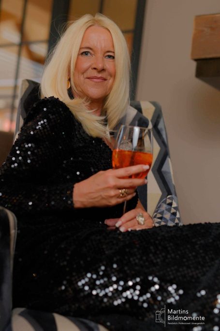 Frau mit blonden Haaren in schwarzem, glitzerndem Kleid hält zwei Gläser in der Hand.