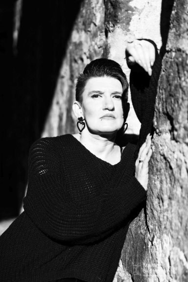 Frau mit kurzen Haaren und auffälligen Ohrringen, lehnt an einer Steinwand.