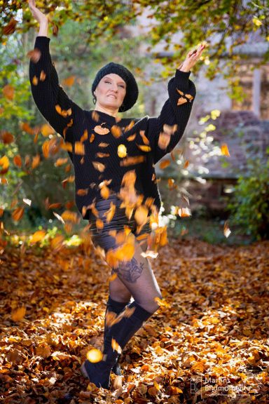 Frau in schwarzer Kleidung, die mit Blättern in einem herbstlichen Park spielt.