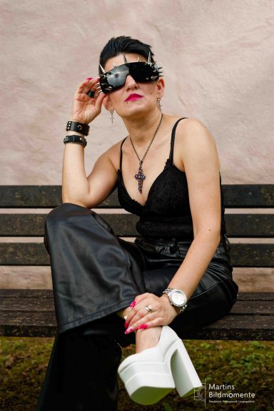 Frau mit Sonnenbrille, schwarzem Outfit und weißen Schuhen sitzt lässig auf einer Bank.
