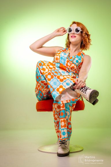 Frau mit rotem Haar in buntem, retro Outfit sitzt auf einem Hocker.