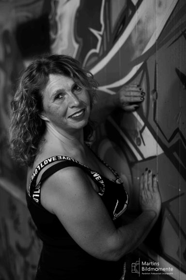 Frau mit lockigem Haar, lächelnd vor einer graffiti-artigen Wand.