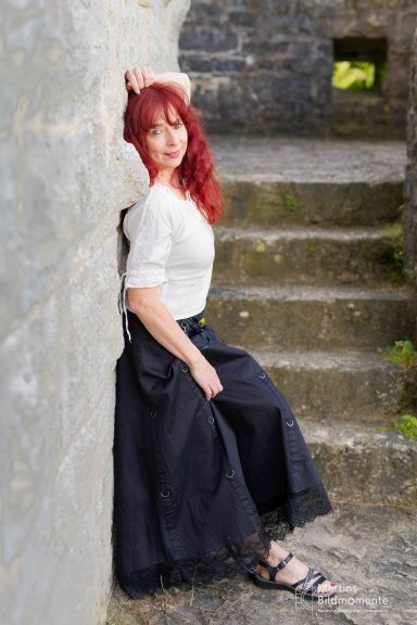 Frau mit langen roten Haaren in Weiß und schwarzem Rock, lehnt an einer Steinmauer.