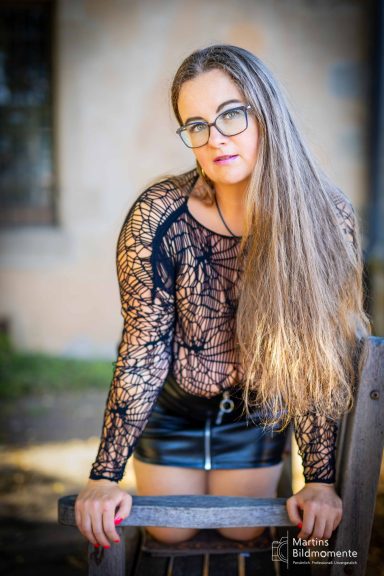 Junge Frau mit langen Haaren, Brille und schwarzer, durchsichtiger Bluse, steht vor einer Wand.