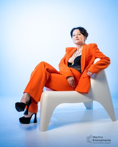 Frau in orange Anzug mit schwarzem Oberteil sitzt auf einem weißen Stuhl.