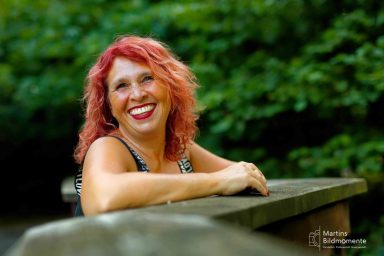 Frau mit rotem Haar lächelt entspannt auf einer Holzbrücke in der Natur.