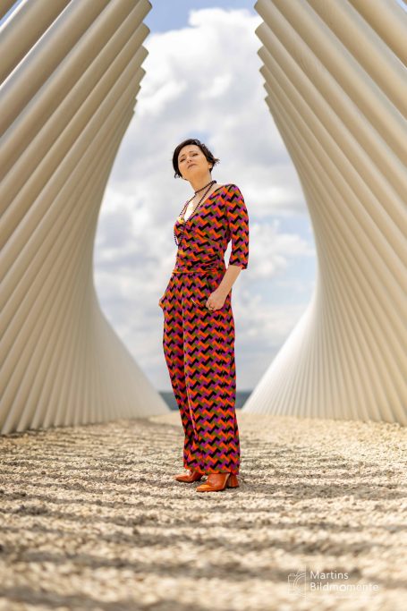 Frau in einem gemusterten Jumpsuit steht zwischen modernen architektonischen Elementen.