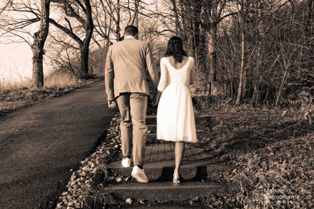 Paar in eleganter Kleidung, Hand in Hand auf einem Waldweg, umgeben von Bäumen.