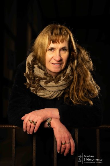 Frau mit langen, blonden Haaren und einem Schal, lächelt in einer dunklen Umgebung.