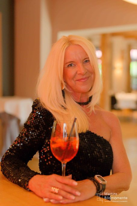Eine blonde Frau in einem eleganten schwarzen Outfit hält ein Getränk in der Hand.
