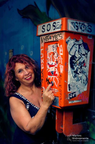Frau mit lockigem, rotem Haar steht neben einem orangefarbenen, graffiti-beschmierten Notrufkasten.