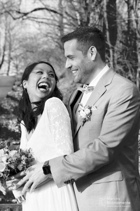 Lächelndes Brautpaar in Hochzeitskleidung, umgeben von Natur, mit einem Blumenstrauß.