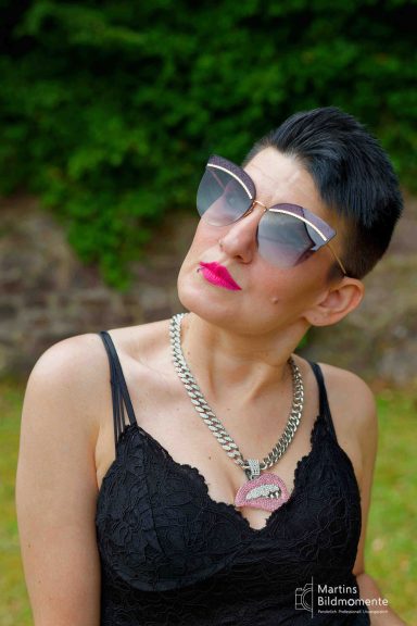 Frau mit kurzen schwarzen Haaren, großen Sonnenbrille und auffälliger Halskette.