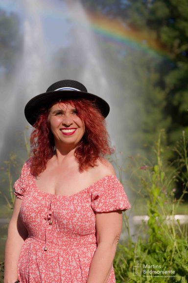 Frau mit roten Locken in einem Blümchenkleid und Hut vor einem Regenbogen.