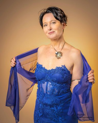Frau in blauer Spitzenkleidung mit offenem Umhang und großen Anhänger um den Hals.