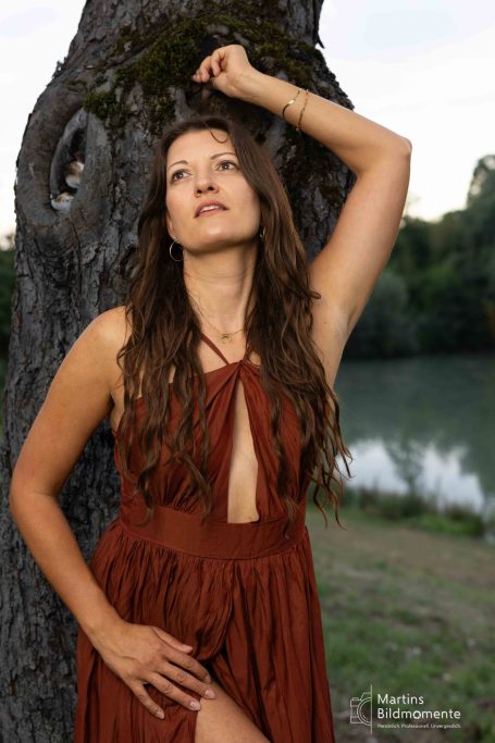 Frau mit langen, braunen Haaren in einem braunen Kleid, lehnt an einem Baum.