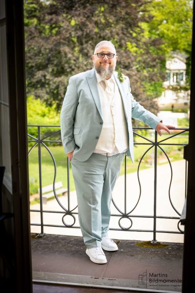 Mann in heller Anzugjacke steht an einem Balkon mit grüner Landschaft im Hintergrund.