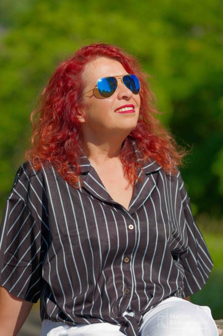 Frau mit roten Locken und Sonnenbrille in einem sonnigen, grünen Umfeld.