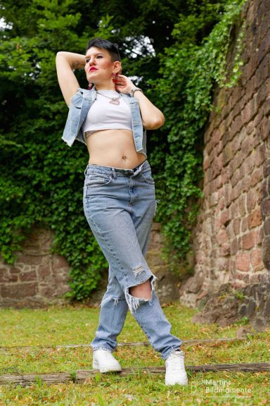 Junge Frau in Jeans und Cropped Top posiert vor einer bewachsenen Mauer.