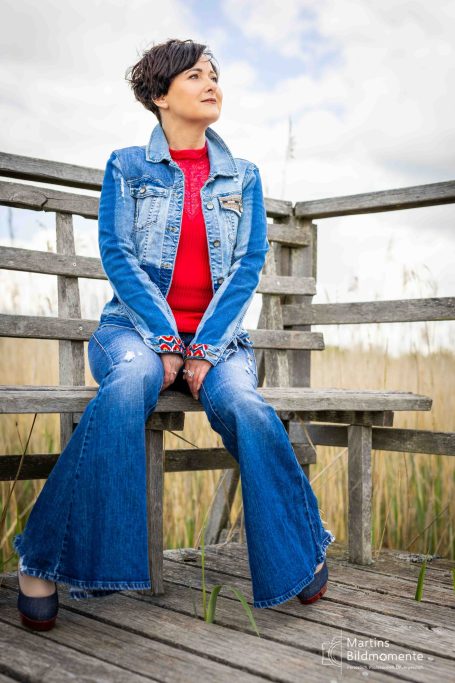 Frau in Jeans und Denimjacke sitzt auf einer Bank in einem Feld mit grauem Himmel.