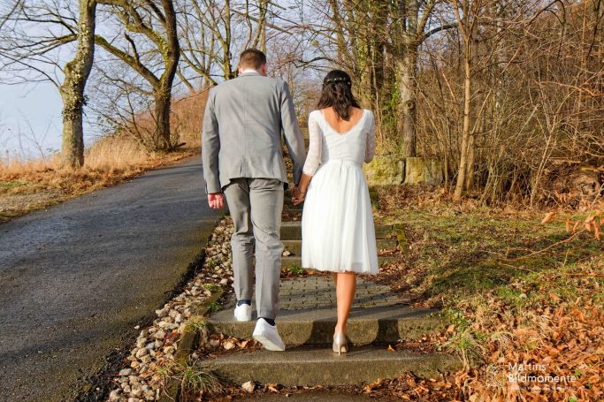 Braut und Bräutigam gehen Hand in Hand einen Waldweg entlang.