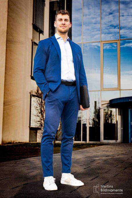 Ein junger Mann in blauem Anzug und weißen Sneakers steht vor einem Gebäude.