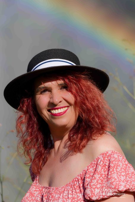 Lächelnde Frau mit rotem Haar und Hut vor einem Regenbogen.