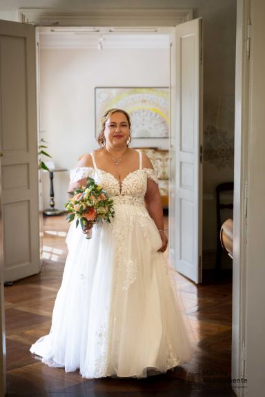 Braut in einem weißen Kleid hält einen Blumenstrauß und steht in einem hellen Raum.