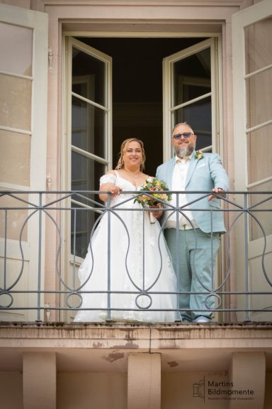 Hochzeitspaar steht auf einem Balkon und lächelt, Braut mit Blumenstrauß in der Hand.