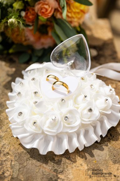Brautpaar-Kissen mit Ringen, weißen Rosen und glänzenden Perlen, auf einer Steinoberfläche.