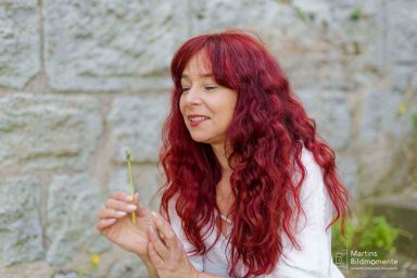 Frau mit langen, lockigen roten Haaren hält eine Blume und lächelt.