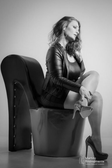 Frau in schwarzem Outfit sitzt auf einem hohen, eleganten Möbelstück und trägt hohe Absätze.