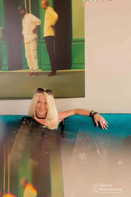Frau mit blonden Haaren sitzt entspannt auf einem Sofa, im Hintergrund zwei Personen.